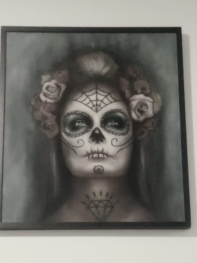A framed portrait depicts a woman with Day of the Dead-style makeup. Her face features a spiderweb design on the forehead and elaborate swirls around the eyes. Roses adorn her hair and a diamond tattoo is visible on her neck. The artwork is monochromatic, with a subtle use of light and shadow enhancing the image's depth.