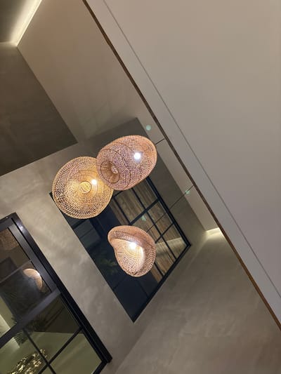 The image displays an upward-angled interior view of a room in Kerobokan Kelod, Indonesia, illuminated by artificial light. Three pendant light fixtures, constructed from woven natural material, are centrally positioned. The largest, disc-shaped fixture is located slightly left of center, emitting a bright, warm glow. A second, similar but smaller disc-shaped fixture hangs above and to its right. A third, irregularly shaped woven pendant light is visible towards the lower right. All lights are illuminated.
The background comprises architectural features. A textured, medium-toned wall occupies the lower-left and central-left portions of the frame. Embedded within this wall are multiple large windows with black frames. These windows reflect interior light and show indistinct, dark exterior views, possibly featuring vegetation. To the upper right, a smooth, light-colored wall meets a ceiling plane, forming a distinct corner. A dark brown trim runs along the intersection of the light-colored wall and the adjacent wall/ceiling. A linear, recessed light source is visible along a ceiling edge in the upper-left area. No individuals or objects indicating human activity are present. The scene is static, under indoor lighting conditions.