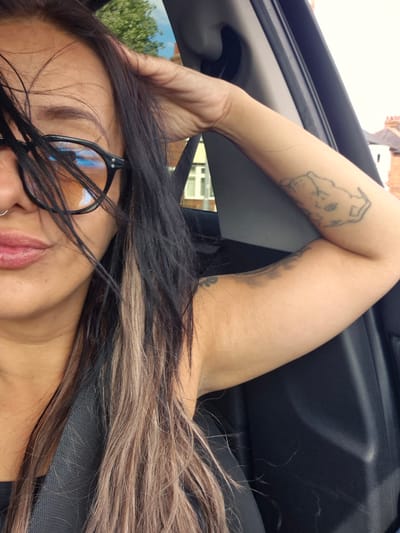 This is a close-up, over-the-shoulder shot of a woman sitting in the passenger seat of a car. She is wearing round, black-rimmed glasses with tinted lenses and has a silver hoop nose ring. Her hair is long and dark with lighter brown and blonde streaks, and it is falling around her face. Her left arm is bent, with her hand resting on her head, and a tattoo of a dog's head is visible on her upper arm. Her right arm is also visible, with a darker, smudged tattoo on her forearm. The interior of the car is dark and blurred, with a glimpse of a building and trees in the background through the window. The scene appears to be a residential street in Bridlington, United Kingdom, given the style of the houses visible in the background.