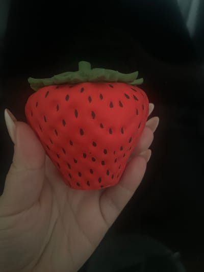 This media file shows a person's hand holding a red, strawberry-shaped object with black seeds. The object has a green, leafy stem. The hand has manicured fingernails that are light pink and appear to be artificial. The background is dark and out of focus. The lighting is dim, suggesting the image was taken indoors in low light conditions. There is no visible text. The scene is very close-up, focusing on the hand and the strawberry object. The context of Torquay, United Kingdom, does not provide specific visual cues within the image itself. The activity appears to be a simple demonstration or display of the object.