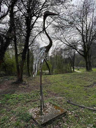 The scene depicts a metallic sculpture of a stylized heron or crane positioned in a park or natural area within Witten, Germany. The sculpture, centrally located, features a dark, curved neck and head, with a body composed of numerous overlapping, weathered light-colored metallic pieces. It is anchored to a rectangular concrete base, partially covered with debris and moss. The immediate ground level is covered with scattered dry leaves and interspersed green growth. Behind the sculpture, a grassy expanse with areas of exposed earth leads to a visible winding path in the midground, which curves into a densely wooded background. The trees are predominantly deciduous and bare of leaves, indicative of late winter or early spring. The overcast sky is visible through the tree canopy. No individuals or dynamic activities are present in the static scene.