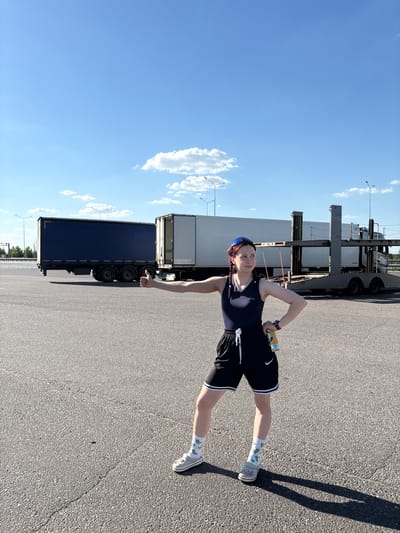 A woman stands on an asphalt surface with a large truck in the background. The woman is wearing a dark tank top, black shorts with white trim, white socks with a pattern, and grey sneakers. She has braided hair and is holding a can in her right hand. A large truck is parked behind her. The sky is blue with some clouds. The location is in Казань, Russia.