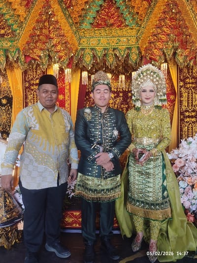 An image captures three individuals standing side-by-side against a richly decorated, festive backdrop, likely at a wedding or formal celebration.

On the left, an adult man is positioned, wearing a long-sleeved collared shirt with a yellow-gold base and intricate light blue and grey batik patterns. He has a black skullcap (peci) on his head, dark trousers, and dark athletic shoes. He is smiling faintly, looking towards the camera.

In the center stands a younger man, dressed in elaborate traditional attire. His outfit consists of a dark green long-sleeved top or jacket heavily embellished with gold embroidery and beadwork, a matching dark green lower garment that appears to be a skirt-like piece with golden fringed trim, and dark green trousers underneath. He wears an ornate golden headpiece. His eyes are closed, perhaps in a blink or a moment of reflection, and his hands are clasped in front of him, with a silver watch visible on his left wrist. He is wearing dark dress shoes or boots.

To the right, a woman is adorned in a stunning, equally elaborate traditional gown. Her dress is primarily green and gold, shimmering with sequins and intricate embroidery, featuring long sleeves and a flowing skirt-like overlay. She wears a magnificent, tall golden headpiece with multiple dangling ornaments, framing her face. She has noticeable makeup and is smiling gently towards the camera. Her hands are delicately posed in front of her, revealing intricate henna (mehndi) designs, and she appears to be holding a small white object, possibly a phone or card. She is also wearing ornate, sparkling shoes that complement her attire.

The background features a vibrant, traditional canopy or archway, adorned with rich red and golden fabrics, intricate patterns, and hanging golden tassels and beadwork. The overall aesthetic suggests an elaborate cultural setting. To the far right, part of a floral arrangement with light-colored flowers is visible, further indicating a celebratory event.

The image is well-lit. A digital timestamp in the bottom right corner reads "05/02/2026 14:37," indicating the photo was taken on February 5, 2026, at 2:37 PM. The scene conveys a celebratory and formal atmosphere.
