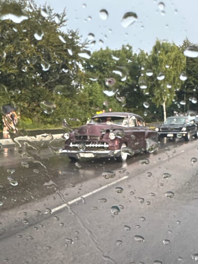 The image shows a vintage burgundy car driving on a wet street, with numerous raindrops obscuring the view. Trees and other cars are visible in the background, suggesting an outdoor setting, possibly in a city. The specific location in Falkenberg, Sweden, is not identifiable from the visual information provided in the image. There is a person walking on the side of the road holding an umbrella. The car appears to be in motion, and the overall atmosphere is one of a rainy day.