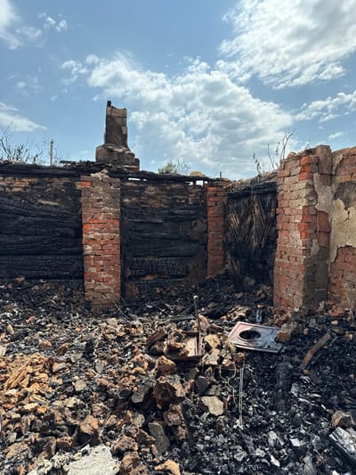 The image displays the fire-damaged interior of a building in the city of Nedryhailiv, Ukraine, under a blue sky with scattered white clouds. The structure's walls are reduced to charred black wooden sections and remnants of red brick pillars, with a damaged brick chimney stack standing centrally against the skyline. The roof is absent. The ground is covered by a dense accumulation of black charred debris, including wood fragments and ash, intermingled with reddish-brown rubble. In the foreground, a cast-iron stove plate with a circular opening and an additional metal object are visible amidst the debris. No people are present within the scene. Sparse, dry vegetation is visible in the background along the perimeter of the destroyed structure.