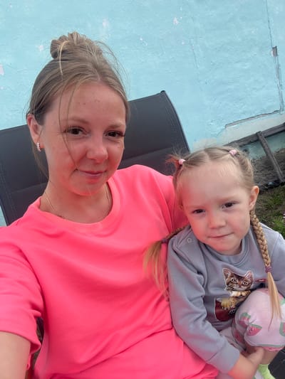 A selfie captures a woman and a young girl sitting outdoors against a light blue, weathered wall. The woman, positioned on the left, is wearing a bright pink long-sleeved shirt and has her hair pulled up in a messy bun. She is smiling and looking towards the camera. The young girl, to the right, is wearing a grey long-sleeved shirt with a cat graphic and pink patterned leggings. Her blonde hair is styled in two pigtails and a braid. She is looking towards the camera with a slight smile. They appear to be sitting in outdoor chairs. The background includes a wooden fence and some overgrown grass. The lighting suggests it is daytime. The location is identified as Сухринское, Russia.