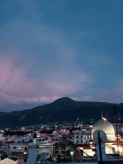 This is a horizontal, medium-shot, eye-level photograph of a city skyline at dusk, featuring a large domed building and mountainous terrain in the background. The sky is a gradient of deep blue to a soft lavender, with scattered clouds. Below the sky, dark mountains are silhouetted against the horizon, with numerous small lights indicating a city nestled at their base. In the foreground, the rooftops of various buildings are visible, with some displaying lit windows. A prominent, pale green domed structure, likely a religious building, is situated in the mid-right of the frame. Several satellite dishes and antenna structures are also visible on the rooftops in the foreground, obscuring some of the urban landscape. The overall impression is of a tranquil evening scene in a coastal city with a mountainous backdrop. The location is the Municipal Unit of Aegio, Greece.