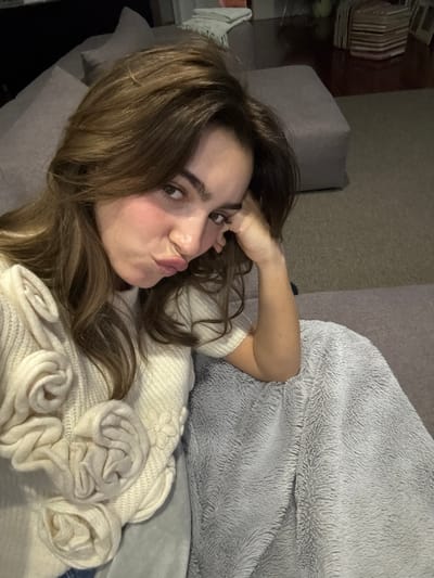 A young woman is depicted in a close-up, slightly overhead shot, lying on a grey sofa in an indoor setting. She has long, wavy dark brown hair and is looking directly at the camera with a playful "kiss face" expression, her lips pursed. One hand is raised, with fingers lightly touching her forehead near her eyebrow.
She is wearing a cream-colored knitted sweater featuring prominent, three-dimensional, rose-like fabric embellishments on the front and sleeves. A fluffy, light grey blanket is draped over a portion of her body and the sofa beside her. The background appears dimly lit, suggesting the photograph was taken in the evening or at night. A neutral-colored wall is visible, along with a glimpse of a table on the far left, where some stacked items (possibly books or decorative boxes) are placed. The scene conveys a relaxed and casual atmosphere. No text is visible in the image. The location context provided is Nogueira e Silva Escura, Portugal.