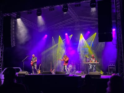 The image depicts a live music performance on an outdoor stage, likely in Tutrakan, Bulgaria, illuminated by purple and yellow stage lights. A band of at least four musicians is performing. In the foreground, a man in a dark shirt and ripped jeans is singing into a microphone, gesturing with his left hand. To his left, another man in a dark shirt and jeans plays a red electric guitar. Further to the right, a man wearing glasses and a dark t-shirt with a graphic print is seated at a keyboard. Behind the singers, a drum kit is visible. Several guitars are propped up on the stage, and large speaker cabinets are positioned on either side of the stage. The audience is partially visible in the foreground, with silhouetted figures watching the performance. The stage structure, including metal trusses and lighting equipment, is prominent above the performers. Smoke or fog is being used to enhance the lighting effects.
