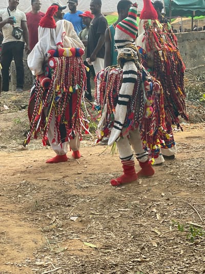 The image portrays an outdoor cultural event in Umueze, Nigeria, featuring three individuals in elaborate masquerade costumes positioned on a dirt ground scattered with dry leaves and debris. In the center and left mid-ground, two primary masquerades are prominently displayed. The figure on the left wears a white flowing garment, a red knitted cap under a white cowl, and is heavily draped with numerous braided red, white, and black ropes adorned with yellow, blue, and green tassels. This figure wears red fabric footwear. The central figure wears a white knitted costume with black horizontal stripes on the upper body and arms, a multi-striped cap (green, red, white, black), and is similarly adorned with braided cords and colorful embellishments, including small multi-colored spheres. This central figure wears red fabric footwear over striped socks. A third masquerade is partially visible on the far right, also featuring braided red, white, and black adornments and a red cap. In the background, approximately eight to ten casually dressed male spectators are observing the event, some smiling, standing behind a low, indistinct barrier. The scene is illuminated by natural, diffused outdoor light.