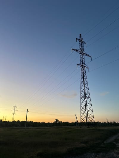 The image displays a landscape scene at dusk in Filipeni, Moldova. The primary subjects are several electricity pylons and the power lines they support, silhouetted against a gradient sky that transitions from a deep blue at the top to a pale yellow and orange near the horizon. The scene is set in a rural or semi-rural environment with a field of grass and a line of trees in the background, partially obscuring what might be a road or pathway on the right side of the frame.

The time of day is clearly sunset or twilight, indicated by the soft, warm colors in the sky. The weather appears clear, with only a few wispy clouds visible. The low angle of the sun casts long shadows and creates a dramatic, atmospheric effect. The electricity pylons are the dominant man-made objects, stretching across the frame and conveying a sense of infrastructure within the natural landscape. The overall mood is one of quiet contemplation, with the fading light and the imposing structures creating a slightly dramatic yet peaceful ambiance. No people are visible in the image. There is no visible text.