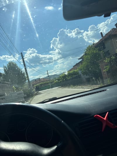 A view from inside a car. The car is stopped on a road in Mikrevo, Bulgaria. A red, four-pointed, star-shaped object is attached to the dashboard near the air vent. The steering wheel and dashboard are visible in the foreground. Through the windshield, a street with houses, trees, utility poles, and a bright blue sky with puffy clouds is visible. The houses appear to be made of brick and have tiled roofs.