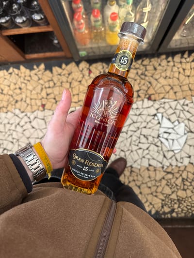 A person's left hand holds a bottle of Havana Club Iconica Gran Reserva Añejo 15 Años Limited Reserve Rum, filled with amber liquid, in an indoor setting within Boulogne-Billancourt, France. The hand exhibits a silver-toned wristwatch on the wrist and a yellow woven bracelet adjacent to it. The individual wears a brown zippered jacket and dark trousers, with a brown shoe visible on the right. The background features a mosaic tiled floor composed of irregularly shaped light-colored tiles, along with a blurred display cabinet containing multiple bottles and a rack of inverted glasses.