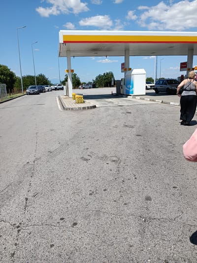 The image shows a gas station with a Shell canopy under a blue sky with white clouds. Several cars are visible, including a gray sedan, a white SUV, and a minivan. A woman wearing a black and white tank top and black pants is walking away from the camera, and a pink object, possibly clothing, is visible in the lower right corner. Two yellow bins are located near the pumps. The ground is asphalt with some cracks and dark stains. Tall light poles are positioned along the road. The scene is likely in Radinovo, Bulgaria, as indicated by the context.