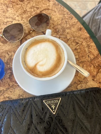 A close-up, overhead shot shows a cappuccino with latte art, a pair of sunglasses, and a black wallet with a "Guess USA" logo on a textured, marble-patterned table. The scene appears to be set in Otranto, Italy, based on the context provided. The cappuccino is in a white cup and saucer, with a spoon resting on the saucer. The sunglasses are gold-rimmed with brown lenses. The wallet is shiny black with a quilted pattern.