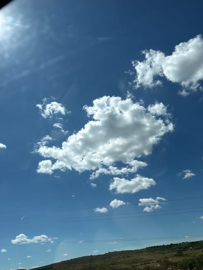 The image shows a bright blue sky filled with fluffy white cumulus clouds. The sun is visible in the upper left corner, creating a lens flare effect and casting light rays across the sky. Below the sky, there is a green, rolling hillside dotted with trees. Several power lines are strung across the lower portion of the sky, running horizontally. The overall scene is a natural landscape with man-made elements. The location is described as Плодовское сельское поселение, Ukraine.