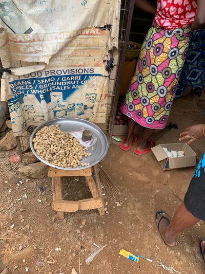 In Mape, Nigeria, this scene captures an outdoor market stall. A large silver tray filled with peanuts is displayed on a wooden stool. Behind the tray, a faded white banner with blue text advertises "FOOD PROVISIONS" and lists various goods such as "BEANS / SEMO / GARRI / YAM / SPICES / GROUNDNUT OIL / PA / FISH / WHOLESALES AND RETAIL." A woman, partially visible, wears a vibrant pink, green, and orange patterned skirt and a red top with white hearts. Her feet are adorned with red flip-flops. To the right, another person's legs are visible, wearing black shorts and black flip-flops. A cardboard box containing several small, white, bagged items sits on the ground near the box. The ground is composed of reddish-brown dirt and scattered stones.