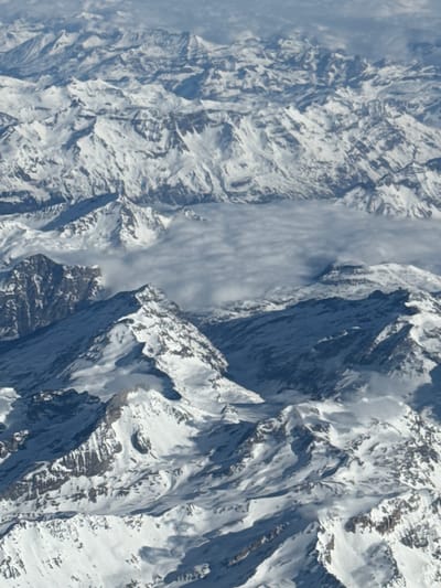 The image presents an aerial perspective of an extensive snow-covered alpine mountain range, characteristic of the region near Cesana Torinese, Italy. Numerous jagged peaks and defined ridges dominate the landscape, primarily blanketed in white snow. Interspersed among the snow are exposed sections of rock, appearing in shades of dark grey and brown, particularly on steep inclines and areas where the snow layer is thinner. A layer of stratiform clouds or fog partially fills the valleys and lower depressions between the mountain formations, creating a soft, diffuse boundary. The scene is illuminated by natural daylight, evident from the shadows cast on opposing mountain faces, indicating depth and topography. No human subjects, artificial structures, or overt signs of activity are visible within the frame, depicting a desolate, high-altitude environment.