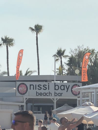 The image captures a lively daytime scene at an outdoor beach bar, likely in Ayia Napa, Cyprus, indicated by the prominent "nissi bay" signage. The clear, bright sky suggests sunny weather.

Several tall palm trees dominate the left and center of the frame, providing a tropical backdrop. On the right, a white structure identifies itself as "nissi bay beach bar" through a large sign featuring the name in black letters and a circular logo with "nb" and smaller text "nissi bay beach." Two orange flags, similarly branded with "nissi bay" and the "nb" logo, are attached to poles near the bar.

Numerous people are visible, primarily on the right side of the image. In the immediate foreground, part of a man's face with glasses is seen. Behind him, a man with a bare chest has his arms raised, engaging in an activity that suggests dancing or stretching. Further back, a crowd, including adults and children, gathers around white beach umbrellas and canopies, indicating a bustling atmosphere of leisure and enjoyment at this popular beachside establishment.