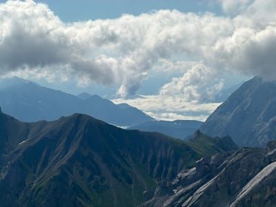 The image depicts a majestic mountain landscape with layered peaks receding into the distance. In the foreground, rugged, dark gray rock faces are sharply defined, with deep crevices and rough textures. Ascending from these rocky slopes are steep, dark green mountainsides, interspersed with patches of exposed dark soil and scree. Some of these slopes are carved with numerous gullies, suggesting erosion. A small patch of white snow or ice is visible on one of the slopes.
The middle ground is dominated by softer, more rounded mountain ranges that appear in shades of blue and gray due to atmospheric perspective. These mountains are less sharply defined than the foreground peaks, with their contours softened by distance and haze.
The background is a layered panorama of further mountain ranges, progressively fading into a pale blue hue. Above the mountains, the sky is a bright blue, dotted with large, billowy white and gray clouds. These clouds are casting shadows on the mountain ranges below, creating a dynamic play of light and shadow across the landscape.
This is a natural mountain scene, likely an alpine environment, with no signs of human presence or urban development visible in the frame. The image captures a sense of vastness and the raw beauty of a rugged mountain terrain under a dynamic sky. This landscape is consistent with the natural environment found in the vicinity of Wildermieming, Austria, known for its mountainous topography.