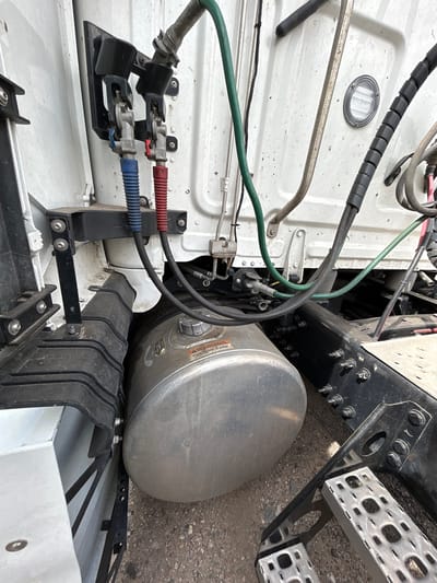 The image depicts the side of a white semi-truck, focusing on its fuel tank and associated components, located outdoors on a gravel surface in Aurora, United States of America. A large, silver, cylindrical fuel tank is prominently visible, positioned horizontally beneath the truck's frame. Attached to the fuel tank is a yellow warning label with red lettering. Above the fuel tank, a black bracket holds two air hose connectors, one with a blue hose and the other with a red hose. Various other hoses and cables, predominantly green, black, and grey, are routed along the truck's chassis. A silver metal step for accessing the truck's cab is situated to the right of the fuel tank. The truck's white body is visible in the background, showing some signs of dirt and dust. The overall environment suggests an industrial or roadside setting.