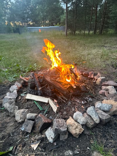 The image depicts a campfire burning in a natural outdoor setting. The fire is in the center of a ring of stones. The ground around the fire is dirt and debris. In the background, there is a grassy area with trees. The lighting suggests it is late afternoon or early evening. There are no people visible in the image. The location is not specified to be in Krutets, Russia.