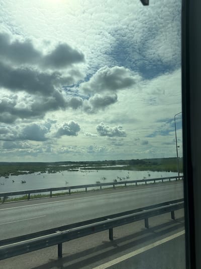 The image depicts a highway scene with a wide, calm body of water adjacent to the road, likely a river or a lake. The water is dotted with green vegetation, appearing to be partially submerged. The sky is filled with dramatic clouds, a mix of dark, heavy storm clouds and lighter, wispy formations, suggesting dynamic weather. A metal guardrail runs along the edge of the highway, separating it from the water. A streetlamp is visible on the right side of the frame, near the horizon. The overall impression is one of a natural landscape viewed from a moving vehicle. Given the provided context, this scene is located in the city of Menzelinsk, Russia.