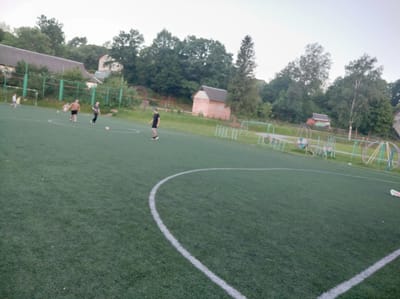 A wide-angle, medium-shot photograph captures a group of individuals engaged in a soccer game on an artificial turf field. The field is marked with white lines, including a curved line in the foreground. In the background, a low, reddish-brown building with a grey roof is visible amongst dense green trees. To the left of the building, a portion of a larger, light-colored structure with a dark roof can be seen. The sky is overcast. Several people are present on the field: on the left, two individuals, one in a yellow shirt and the other in a light-colored shirt, are near a soccer goal. Further into the field, a person in a black shirt is running, and another person is kicking a soccer ball. To the right, there are playground-like structures with colorful elements, including what appears to be part of a merry-go-round. The overall scene depicts a casual sporting activity in an outdoor recreational area.