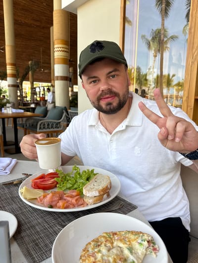 A man in a green hat and white polo shirt sits at a table. He holds up two fingers in a peace sign and a cup of coffee. Two plates with food are visible: one with bread, lettuce, salmon, tomatoes, and cheese, and the other with an omelet. Palm trees are visible in the background through a window.