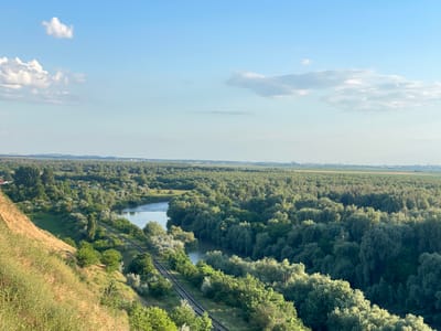 The image shows a panoramic view of a river winding through a dense forest. The river has calm, reflective waters. A set of railway tracks runs parallel to the river, curving along the landscape. The foreground features a steep, grassy slope covered in dry vegetation, with scattered green bushes and trees. The background reveals a vast expanse of lush green trees, stretching towards a distant horizon where a hazy cityscape or industrial area is visible. The sky is a clear blue, with a few scattered white clouds. The scene appears to be captured from an elevated vantage point, looking down and across the river valley. This view is of the Prut River in the vicinity of Cîșlița-Prut, Moldova, a region characterized by its extensive riparian forests and agricultural plains.