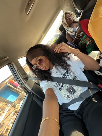 Two women are riding in a car. The woman in the foreground wears a white t-shirt with a graphic print, black pants, and a seatbelt. She also has sunglasses and jewelry on her head and wrist. The woman in the background wears a patterned headscarf and seatbelt. Both women appear to be of African descent. A partial view of the outside shows buildings and possibly market stalls. The image is taken from a slightly low angle within the vehicle.