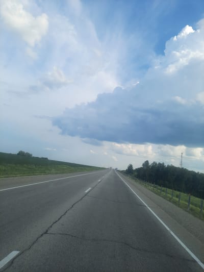 The image shows a view from a moving vehicle on a highway in Oktyabr'skii, Russia. The perspective is from the passenger seat, looking forward and slightly to the right. The road is paved with asphalt and has a white line marking the lanes. The highway is bordered by a grassy shoulder and a fence. To the right of the fence, there is a dense line of trees. The sky is partly cloudy with large, dramatic clouds. The sun is not directly visible, but the sky is bright, suggesting it is daytime, likely mid-afternoon. There is a visible power pylon in the distance on the right side of the frame, behind the trees. The scene conveys a sense of travel and open space. No people are visible, and there is no visible text.