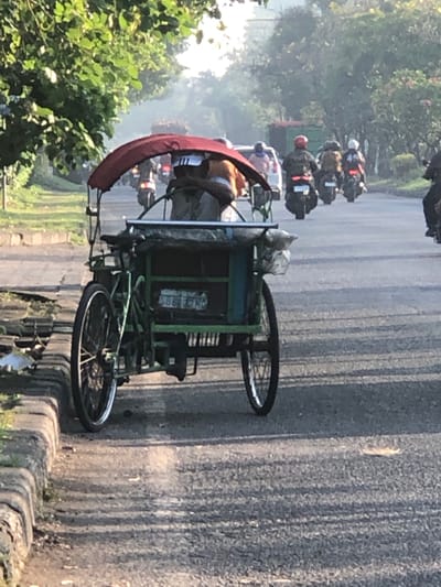 In the early morning light of Ngampelsari, Indonesia, a three-wheeled pedal rickshaw, painted green, is stationary on the right side of a paved road. A person is seated inside the rickshaw, partially obscured by its red canopy. The rickshaw has a bicycle-style front wheel and two wheels at the rear. The road is lined with a concrete curb and vegetation on the left. In the background, several motorcycles with riders are traveling away from the camera, some carrying passengers. Trees line the right side of the road, and a light haze or fog is visible in the distance, typical of early morning conditions. The scene depicts a common mode of transportation in Indonesia, with the rickshaw waiting for passengers or being parked.