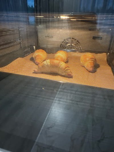 The image shows three golden-brown bread rolls baking on a baking sheet inside an oven. The oven door has horizontal lines, distorting the view of the rolls. The rolls are positioned in the center of the frame, with the baking sheet beneath them. The background of the oven is dark, with a hint of light from the oven's interior light. The foreground shows the oven door, which is reflective and dark. The image does not provide any information about the location in the city of Murom, Russia.