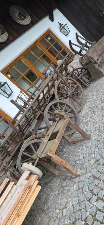 The image shows a collection of antique wooden carts and equipment displayed on a cobblestone surface in front of a building with a white facade and wooden-framed windows. The scene appears to be outdoors, possibly in a courtyard or historical area. The objects are weathered and show signs of age, with one cart prominently featuring large wooden spoked wheels. Some decorative elements, including framed artwork and a lantern, are visible on the building. The overall impression is one of historical preservation and display of vintage agricultural or transport machinery.
