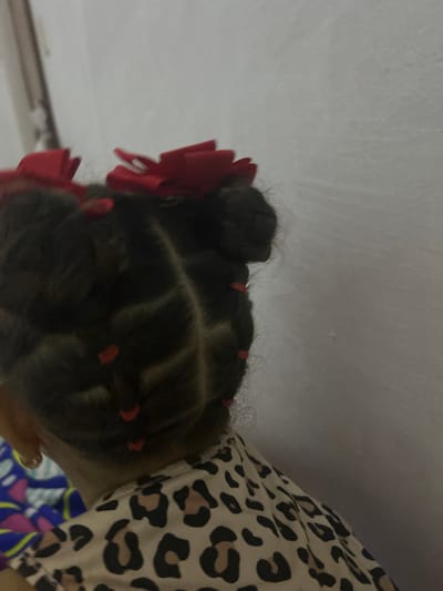 The image shows the back of a child's head with dark hair styled in small buns or sections separated by parts. Small red hair ties or bands secure the sections. A large red bow is attached to one side. The child is wearing an animal-print patterned top. The background is a white wall and part of what appears to be a table. The location is indeterminate, but the request specified San Juan de Los Morros, Venezuela.