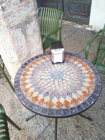 The image shows an outdoor cafe scene in Besalú, Spain. A circular mosaic table with a geometric pattern of brown, gray, and cream tiles is the central focus. On the table sits a silver napkin dispenser with white napkins. Around the table are several green metal chairs. In the background, a rough-textured stone wall and a weathered, dark wooden door are visible. The ground is paved with rough, gray stones. The lighting suggests it is daytime.