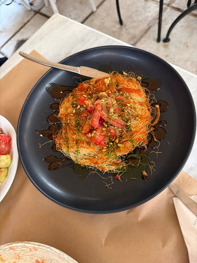 A top-down, medium shot captures a black plate holding a nest-like dessert, possibly Kunafa, topped with chopped tomatoes and pistachios, drizzled with a dark syrup. The dessert sits on a textured brown paper placemat, with a knife resting on the upper left edge of the plate. To the left of the plate, a white bowl contains sliced cucumbers and tomatoes. The background shows a light-colored table with a tiled floor and dark metal chair legs, suggesting an outdoor dining setting in Thessaloniki, Greece.