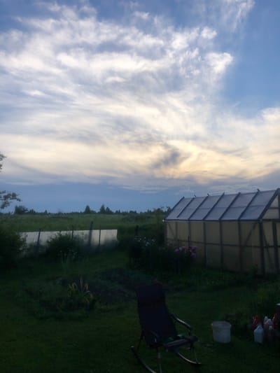 In the city of Uriupino, Russia, a scene unfolds at dusk with a wide, cloudy sky. In the foreground, a dark, weathered rocking chair sits on a grassy lawn, positioned to face a small, weathered greenhouse. The greenhouse, constructed of wood and translucent material, stands near a cluster of pink flowers. Beyond the greenhouse, a vast, green field stretches to the horizon, dotted with trees. A makeshift fence, likely made of wood and some form of plastic sheeting, runs along the edge of the field. The overall atmosphere is serene, with the fading light casting a soft glow over the landscape. There are no people or animals visible in the image.