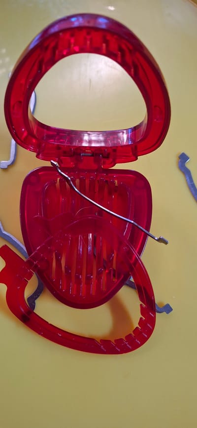 A red plastic hair clip is open, showing the combs inside and a thin metal tool. The clip is heart-shaped with a round handle. Some silver metal objects, possibly hair clips or hairpins, are on the yellow background. The scene is indoors, and the background appears to be a tabletop. The image is a close-up, taken from a high angle.