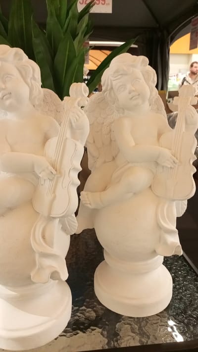 Two white cherub statues with violins sit on a glass surface indoors. The statues are placed next to each other. A man with a beard is visible in the background. The photograph was taken in Yudino, Russia.