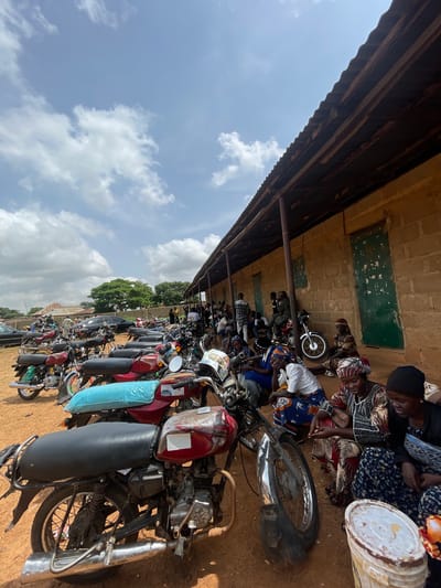 In Kassa, Nigeria, a group of people are gathered outdoors, near a long, single-story building with a corrugated metal roof. The building appears to be constructed from rough, tan-colored bricks. In front of the building, many motorcycles are parked in rows. The ground is reddish-brown dirt. Several people, including women wearing headscarves and patterned clothing, are sitting or kneeling on the ground in front of the motorcycles and the building, some appearing to be conversing or observing their surroundings. Others stand in small groups further back. A black car is visible in the background, as are trees and other structures. The sky is partly cloudy, with white clouds against a pale blue backdrop.