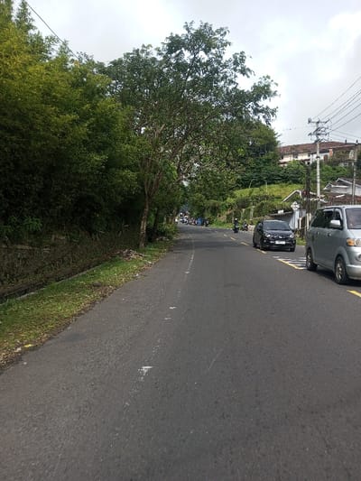 This image depicts a grey asphalt road in Saing Ta Mawt Lay, Myanmar, viewed from a low angle. The road curves slightly to the left in the distance. On the immediate left, a narrow grassy shoulder borders a concrete drainage ditch, beyond which a dense wall of green bamboo and leafy trees extends upwards. On the right, a silver minivan is partially visible, parked or moving slowly. Further down the right lane, a black hatchback is in motion. In the mid-distance on the road, multiple motorcycles, some with riders, are visible, with a cluster of stopped or slow-moving motorcycles and potentially pedestrians on the left side of the road. Uphill on the right, residential structures are visible, including a multi-story building with a red-tiled roof. Utility poles with overhead lines are present on the right. The sky is overcast and light grey. Road markings include faint white dots on the left and solid yellow lines on the right.