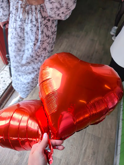 The image shows two inflated, red heart-shaped foil balloons being held by a light-skinned hand with manicured nails and a ring. The background shows parts of three people: one in a long white dress with a dark floral pattern, another in a white top and black pants with white sneakers, and another mostly cut off. The setting appears to be indoors, with a light-colored floor. The location in Gremyachinsk, Russia is not discernible from the visual information provided in the image.