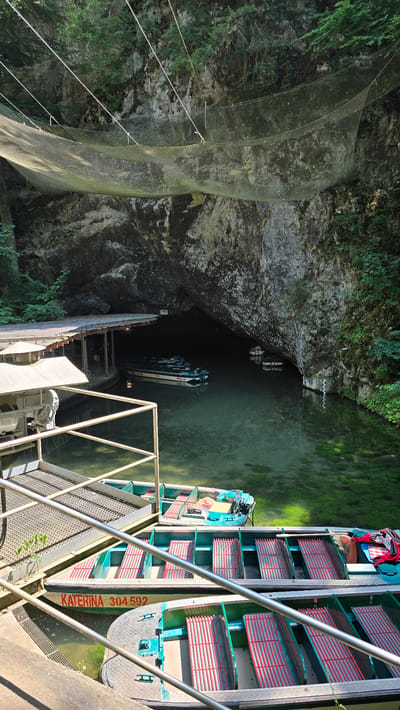 The image depicts the entrance to a cave with a body of water, likely a river or underground lake, and several boats. A covered dock is visible on the left side of the cave entrance, with a few boats moored. The water is a clear, greenish hue, with aquatic plants visible beneath the surface. Further into the cave, more boats can be seen, suggesting this is a popular tourist or transport location. The cave opening is large and rugged, with vegetation growing on the surrounding rock faces. A large net structure is suspended across the upper portion of the cave entrance, possibly for safety or to prevent debris from falling. In the foreground, several boats with rows of red seating are docked. One boat has the name "KATERINA 304592" painted on its side. The overall scene suggests a natural attraction that has been adapted for public access. This location is identified as the Macocha Abyss in the Moravian Karst region of the Czech Republic, near the town of Nové Dvory.