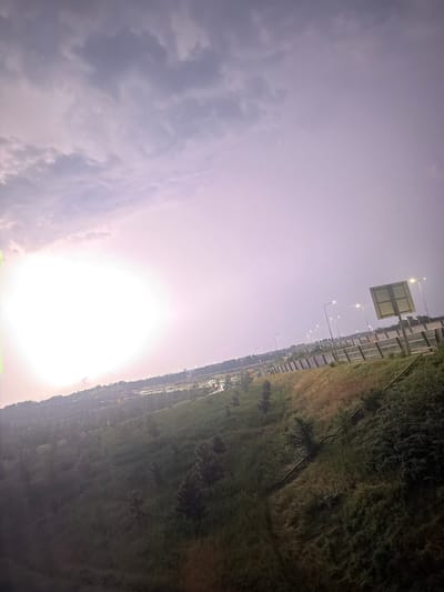 The image captures a scene from Badhoevedorp, Netherlands, likely near a highway or major road. The perspective is from an elevated position looking down and across a sloping grassy embankment. The sky dominates the upper portion of the image, displaying a gradient of soft purple and pink hues, indicative of either dawn or dusk. Large, diffused clouds are present.
In the mid-ground, a highway or roadway is visible, lined with streetlights that are illuminated. Several vehicles are present on the road, their lights suggesting some traffic. A tall signpost with a rectangular sign is positioned on the right side of the road. The embankment is covered with a mix of green and dry grass, with scattered trees and bushes. A set of stairs or a pathway is visible winding down the embankment on the right side of the image.
In the distance, the urban landscape of Badhoevedorp is suggested by buildings and more distant lights, though details are obscured by haze or distance. The overall mood is serene, with the soft lighting and natural elements contrasting with the man-made infrastructure. There are no people visible in the frame. The environment appears to be an edge of a city, where nature meets urban development.