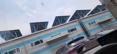 The image depicts the exterior of a modern building with a corrugated metal facade, featuring horizontal blue and silver stripes. The building's roof is lined with multiple solar panels arranged in angled rows, tilted towards the sky. Several windows are visible on the building's exterior, some of which are rectangular and others are long and narrow. In the foreground, the tops of several parked vehicles are visible, including a dark-colored SUV and another dark car. The scene is set against a bright sky with scattered clouds. The overall impression is one of an industrial or commercial building, possibly with a focus on renewable energy.