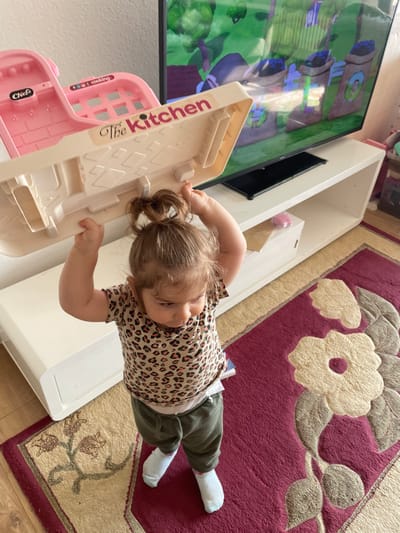 This image depicts an indoor residential scene in Senden, Germany. A young child, with light brown hair styled in a bun, stands on a burgundy-colored rug adorned with large beige floral patterns. The child is wearing a short-sleeved leopard-print shirt, olive green sweatpants, and white socks. She holds a beige and pink plastic toy, bearing the label "The Kitchen" and additional text like "Chef" and "cooking," with both hands raised above her head. In the background, a large flat-screen television rests on a white entertainment console with open shelving, displaying an animated scene featuring green trees and brown sacks containing blue items. The floor surrounding the rug is a light-toned surface.