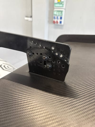 A close-up shot showcases a black metal bracket attached to a surface with a distinctive carbon fiber pattern. The bracket features multiple adjustment holes, labeled with numbers ranging from 4 to 14. Two screws secure the bracket to the surface. The scene is well-lit, highlighting the textures of both the bracket and the carbon fiber. A blurry object and an electrical panel are in the background.