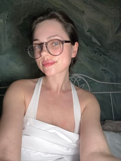 This image captures a close-up of a female subject in an indoor bedroom setting located in Scafati, Italy. The subject, of fair complexion, appears to be an adult, positioned centrally and looking directly into the camera. She has dark hair, light-colored eyes, and wears round, clear-framed prescription glasses. Her lips are full and exhibit a reddish tint, with a small, localized mark present on the upper lip, slightly right of center. She is wearing a white halter-neck garment and is partially covered from the chest down by white bedding or fabric. Behind the subject, a section of a white, ornate metal bed headboard is visible to the right. The wall forming the background features a distinct dark green marbled pattern, characterized by swirling, abstract lines. The scene is illuminated by soft, even lighting.